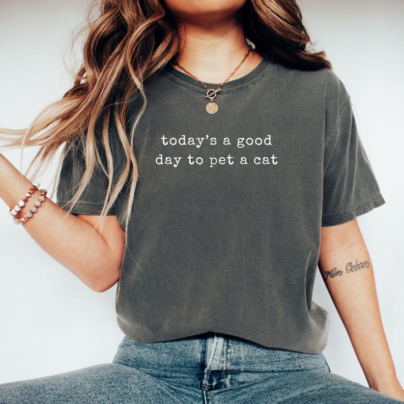 Op de afbeelding: Donkergrijs T-shirt met de witte tekst "today's a good day to pet a cat". Het shirt heeft een ronde hals en korte mouwen. Het model draagt een blauwe spijkerbroek en een gouden ketting.