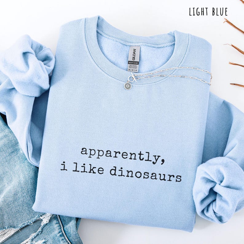 May include: Light blue crewneck sweatshirt with the text "apparently, i like dinosaurs" in black. A silver necklace with a small pendant is on the shirt. The sweatshirt is folded with a pair of blue jeans.