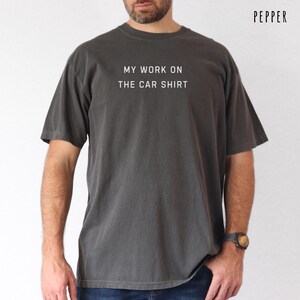 Pepper gray car shirt featuring the phrase My Work On The Car Shirt printed in white lettering. Funny car guy t shirt perfect for mechanic dads car lover husbands auto repair guys garage dads classic car fans weekend wrench turners DIY mechanics hot rod lovers and men who spend their free time working on cars. Great casual automotive gift tee.