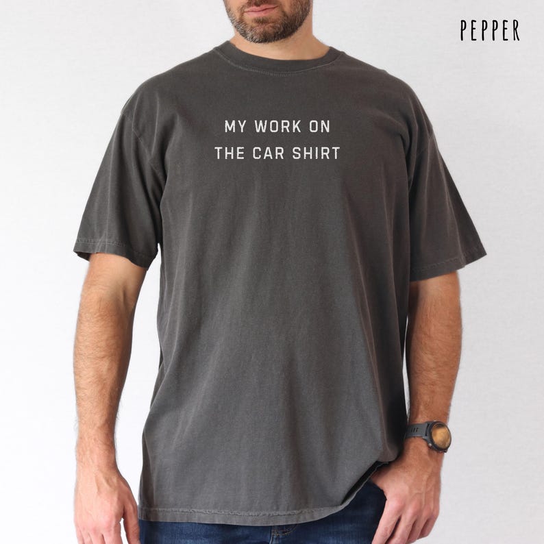 Pepper gray car shirt featuring the phrase My Work On The Car Shirt printed in white lettering. Funny car guy t shirt perfect for mechanic dads car lover husbands auto repair guys garage dads classic car fans weekend wrench turners DIY mechanics hot rod lovers and men who spend their free time working on cars. Great casual automotive gift tee.