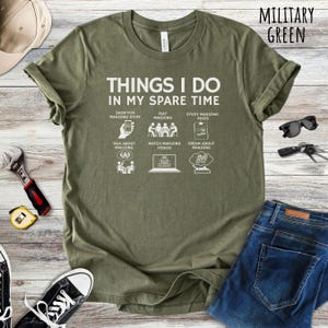 May include: Military green t-shirt with the text "THINGS I DO IN MY SPARE TIME" and illustrations of Mahjong-related activities. The shirt includes the text "SHOP FOR MAHJONG STUFF", "PLAY MAHJONG", "STUDY MAHJONG RULES", "TALK ABOUT MAHJONG", "WATCH MAHJONG VIDEOS", and "DREAM ABOUT MAHJONG".