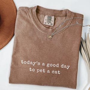 Op de afbeelding: Een bruin Comfort Colors t-shirt met de tekst "today's a good day to pet a cat" in het wit. Een zilveren ketting met een hanger ligt op het shirt. Een bruine hoed en gedroogde palmbladeren zijn op de achtergrond.