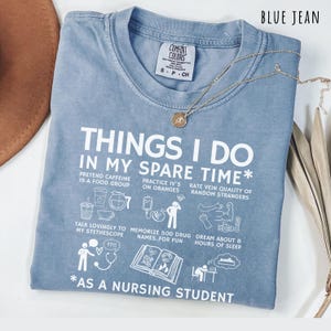 Peut inclure: T-shirt bleu jean Comfort Colors avec texte et illustrations blanches. Le t-shirt indique "THINGS I DO IN MY SPARE TIME* AS A NURSING STUDENT". Un collier dor&eacute; avec un petit pendentif est pos&eacute; sur le t-shirt.