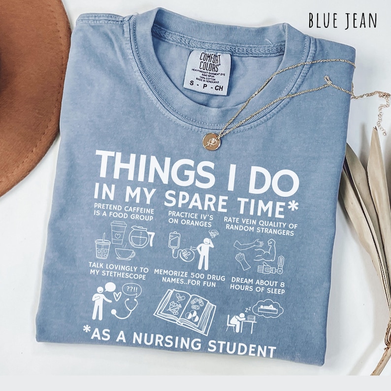 Peut inclure: T-shirt bleu jean Comfort Colors avec texte et illustrations blanches. Le t-shirt indique "THINGS I DO IN MY SPARE TIME* AS A NURSING STUDENT". Un collier dor&eacute; avec un petit pendentif est pos&eacute; sur le t-shirt.