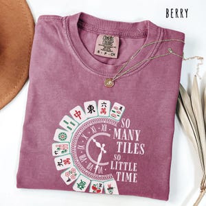 May include: Berry-colored t-shirt with a round graphic featuring Mahjong tiles and the text "SO MANY TILES SO LITTLE TIME." The shirt has a crew neck and short sleeves. A gold necklace with a pendant is draped on the shirt.