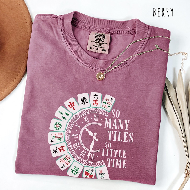 May include: Berry-colored t-shirt with a round graphic featuring Mahjong tiles and the text "SO MANY TILES SO LITTLE TIME." The shirt has a crew neck and short sleeves. A gold necklace with a pendant is draped on the shirt.