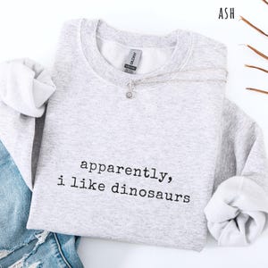 May include: A light gray sweatshirt with the text "apparently, i like dinosaurs" in black. The sweatshirt has long sleeves and a crew neck. A silver necklace with a small pendant is draped across the front. The word "ASH" is in the top right corner.