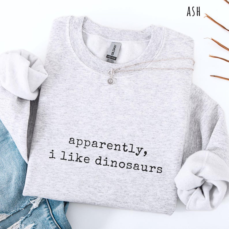 May include: A light gray sweatshirt with the text "apparently, i like dinosaurs" in black. The sweatshirt has long sleeves and a crew neck. A silver necklace with a small pendant is draped across the front. The word "ASH" is in the top right corner.