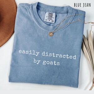 May include: A blue jean colored t-shirt with the white text easily distracted by goats printed on the front. The shirt features a crew neckline and a Comfort Colors brand label at the inner collar. A gold chain necklace with a small round pendant rests on the fabric.