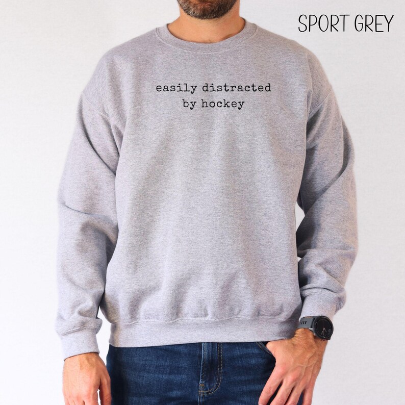 May include: A sport grey crewneck sweatshirt featuring the black text "easily distracted by hockey" printed on the chest. The garment has long sleeves, a ribbed neckline, and ribbed cuffs, providing a standard fit for casual wear.