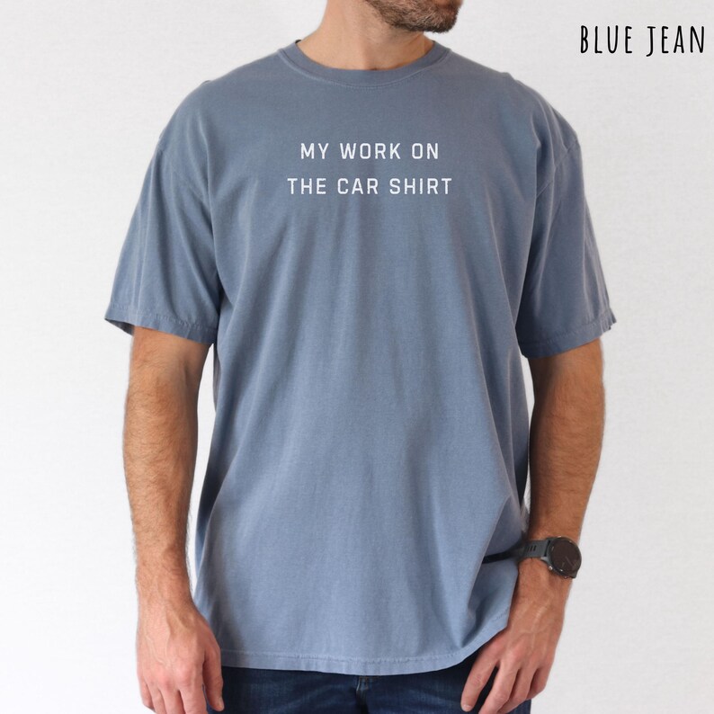 May include: A blue t-shirt with the text "MY WORK ON THE CAR SHIRT" in white. The shirt is a solid color and has short sleeves. The words "BLUE JEAN" are in the upper right corner.