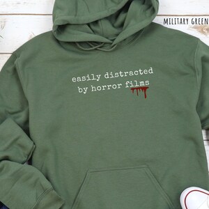 May include: Military green hoodie with the text "easily distracted by horror films" in white, and a red blood drip graphic. The hoodie has a hood and a front pocket.