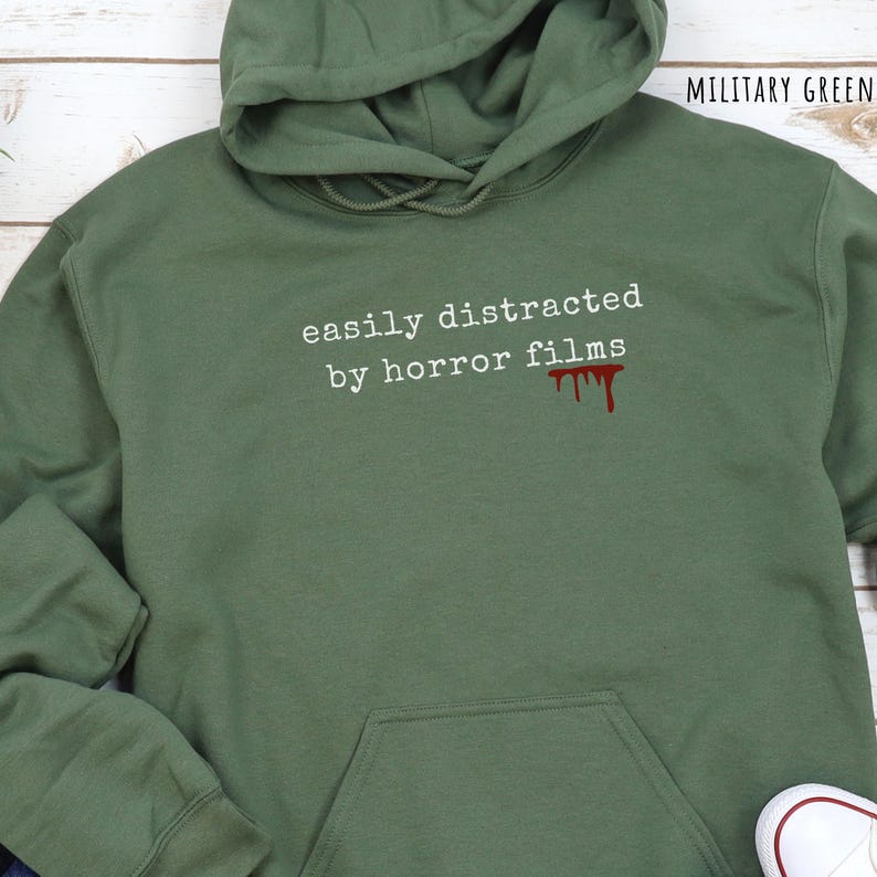 May include: Military green hoodie with the text "easily distracted by horror films" in white, and a red blood drip graphic. The hoodie has a hood and a front pocket.