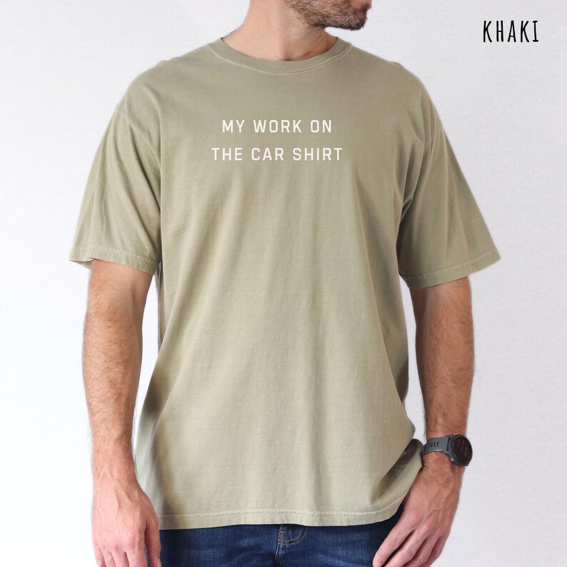 May include: Khaki-colored t-shirt with the text "MY WORK ON THE CAR SHIRT" in white. The shirt is a short-sleeved, crew neck style. The word "KHAKI" is printed in the upper right corner.