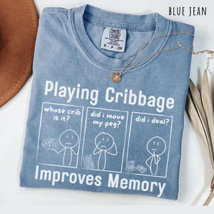 Może przedstawiać: Niebieska koszulka w kolorze jeansu z białym nadrukiem graficznym i napisem Playing Cribbage Improves Memory. Wzór zawiera trzy panele z postaciami z kresek pytającymi whose crib is it, did i move my peg oraz did i deal.