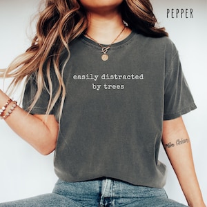 May include: A dark gray t-shirt with the text "easily distracted by trees" in white. The shirt is a crew neck style and appears to be made of a soft material. The word "PEPPER" is printed in the upper right corner.