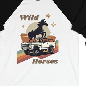 May include: A white and black baseball tee shirt with a retro design featuring a black horse standing on a vintage white Bronco. The design includes a sunset, stars, and the text "Wild Horses".