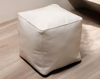 Op de afbeelding: Een witte vierkante poef van leer. De poef staat op een lichte houten vloer.