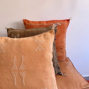 May include: Three decorative throw pillows with embroidered geometric patterns. The pillows are layered on top of each other, with a brown pillow on top of a dark orange pillow, and a light orange pillow on the bottom.