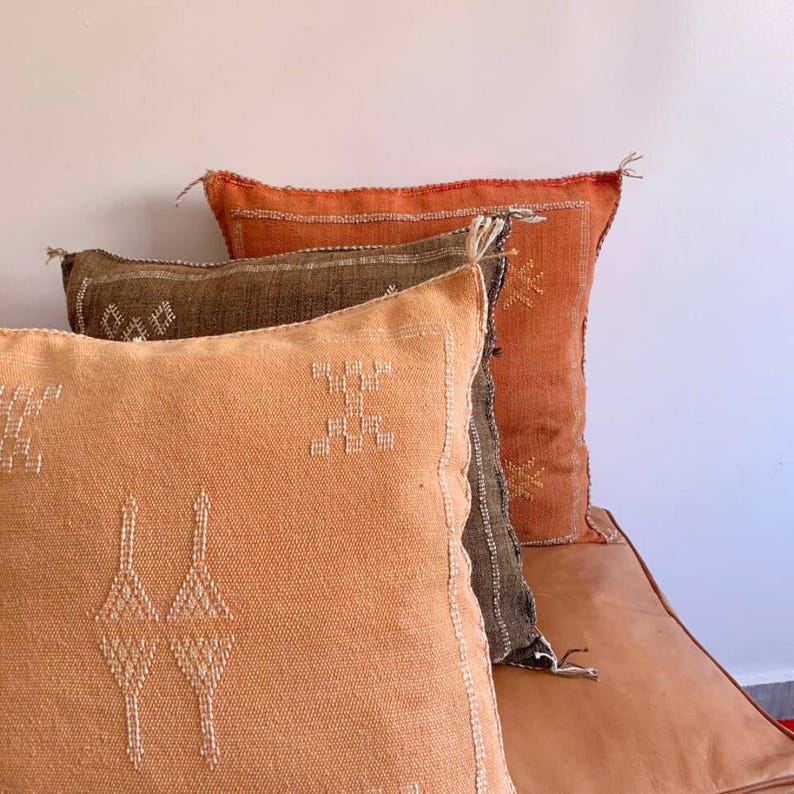 May include: Three decorative throw pillows with embroidered geometric patterns. The pillows are layered on top of each other, with a brown pillow on top of a dark orange pillow, and a light orange pillow on the bottom.
