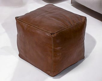 May include: A square, brown leather ottoman. The cube-shaped furniture piece has visible stitching along the seams and a slightly textured surface. The ottoman is a warm, medium brown color and sits on a white surface.