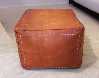 May include: A brown leather ottoman with white stitching. The ottoman is square shaped and has a smooth, textured surface.