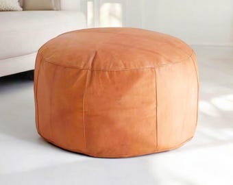 Può includere: Un pouf rotondo in pelle color cuoio con una superficie liscia e leggermente strutturata.