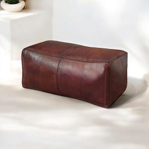 May include: A brown leather ottoman with a rectangular shape and stitched seams. The ottoman is made of genuine leather and has a rustic, vintage look.