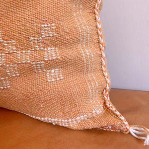 May include: A close-up of a peach-colored pillow with a white geometric pattern. The pillow has a white fringe trim and a white tassel.