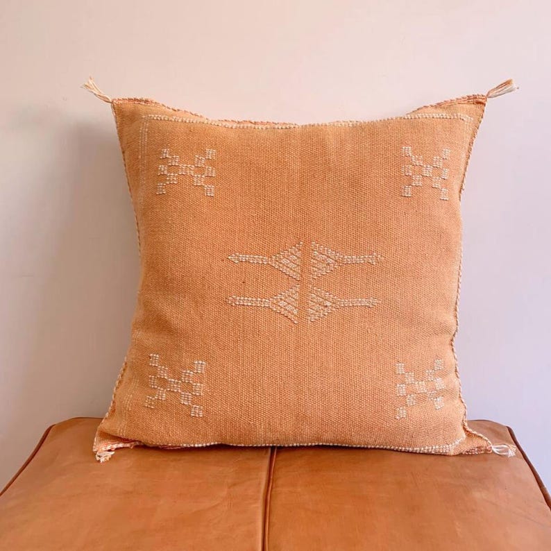 May include: A square, peach-colored throw pillow with a white embroidered geometric pattern. The pillow is made of a soft, woven fabric and has a tassel on each corner.