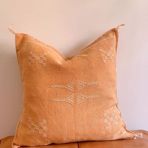 May include: A square, orange throw pillow with a white embroidered geometric pattern. The pillow is made of a soft, woven fabric.