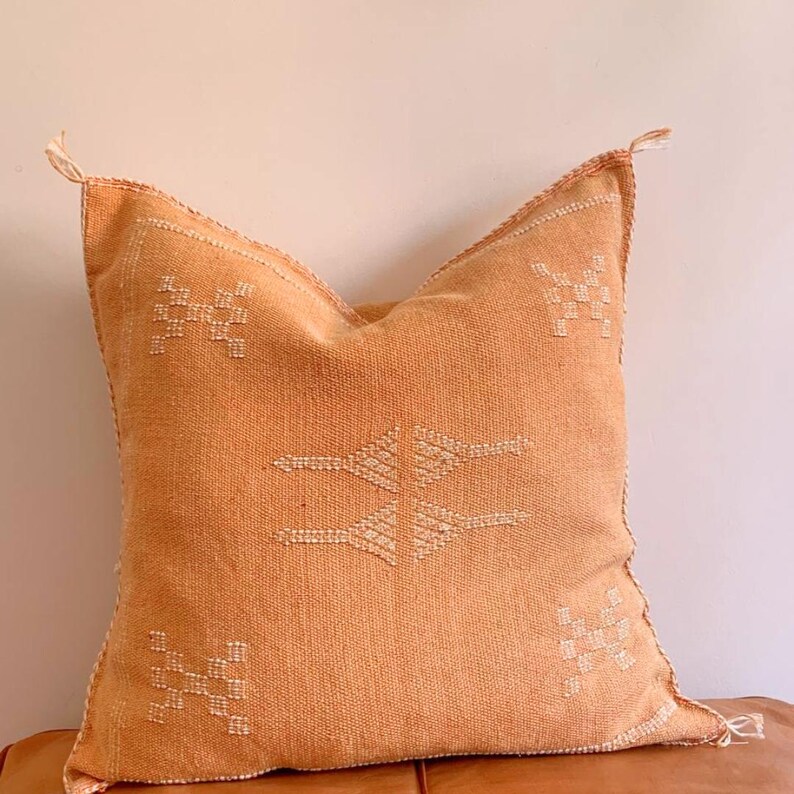 May include: A square, orange throw pillow with a white embroidered geometric pattern. The pillow is made of a soft, woven fabric.