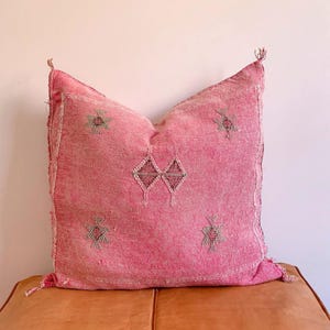 May include: A pink throw pillow featuring a traditional Moroccan design. The pillow has a diamond pattern in the centre and stars on the corners. The pillow is made from a soft, woven fabric.