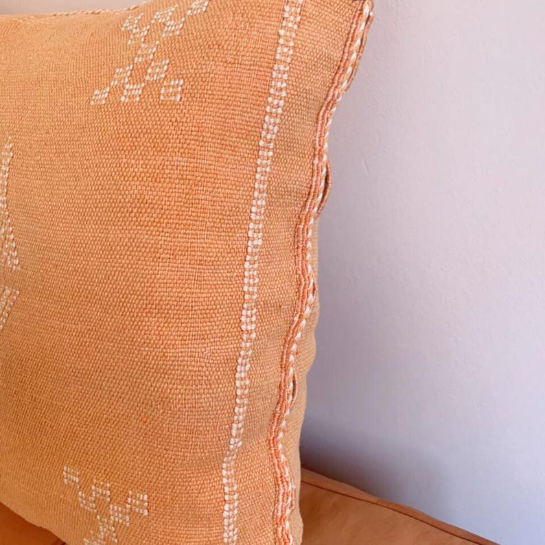 May include: A square, peach-colored throw pillow with white embroidered details. The pillow has a woven texture and a white border.
