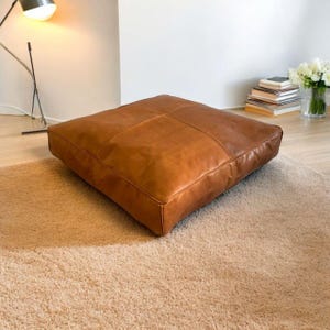 May include: A large, square, brown leather floor cushion. The cushion is made of thick, supple leather and has a simple, elegant design.