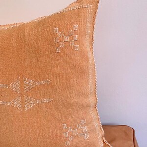 May include: A light orange throw pillow with white embroidered geometric patterns. The pillow has a fringed edge.