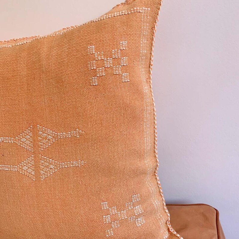 May include: A light orange throw pillow with white embroidered geometric patterns. The pillow has a fringed edge.