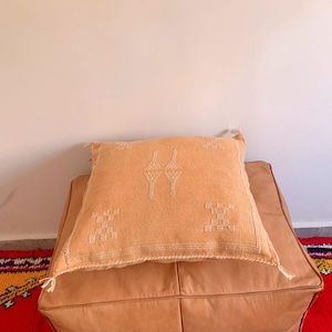 May include: A light orange throw pillow with a white embroidered geometric pattern sits on a tan leather ottoman.