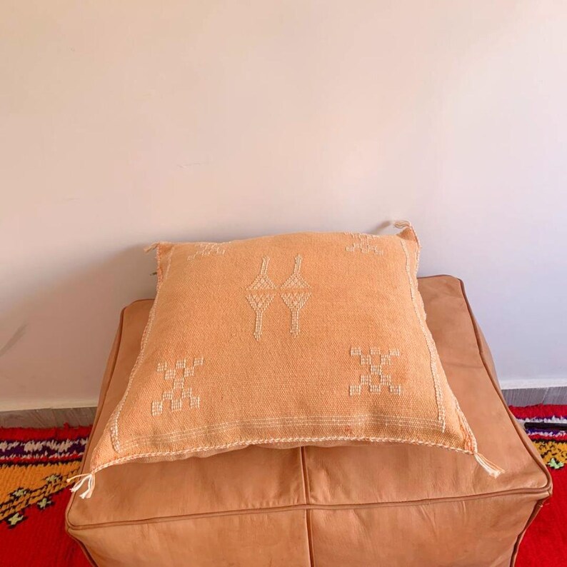 May include: A light orange throw pillow with a white embroidered geometric pattern sits on a tan leather ottoman.