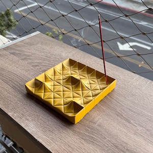 Op de afbeelding: Een vierkante mosterdgele wierookhouder met een geometrisch, gewatteerd oppervlak met verhoogde driehoekige secties. Een enkel dun rood wierookstokje is in de houder geplaatst en verspreidt een dunne sliert witte rook.