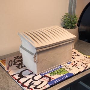 Op de afbeelding: Een kleine, gebroken witte vuilnisbak met een geribbeld deksel. De kleine container staat op een kleurrijke mat met graffiti-achtige ontwerpen en tekst. Een kleine potplant en een spiegel staan op de achtergrond.