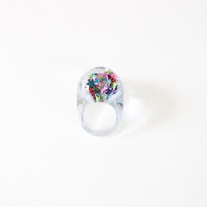 Puede incluir: Un anillo de resina transparente y redondeado que presenta una colección de purpurina en forma de estrella multicolor incrustada en la parte superior. El anillo tiene una banda lisa y transparente y una parte superior en forma de cúpula llena de estrellas rojas, azules, verdes y moradas.