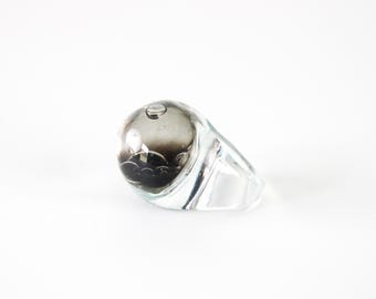 May include: A clear glass ring with a rounded, smoky grey centre. The ring has a smooth, polished finish and a modern, minimalist design. The centre of the ring appears to have a dark, abstract design within the glass.