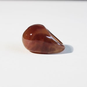 Peut inclure: Une pierre lisse en forme de goutte, de couleur ambre, avec une finition brillante. La pierre présente un aspect moucheté et scintillant, sur fond blanc. L'objet semble être un objet décoratif ou un bijou.