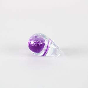 May include: A clear glass ring with a teardrop-shaped design. The ring contains a vibrant purple liquid with bubbles. A thin purple stripe accents the clear glass, creating a unique and artistic piece of jewelry.