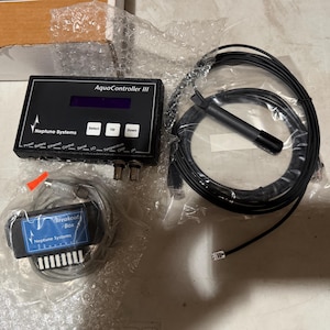May include: A black Neptune Systems AquaController III unit with a digital display and control buttons, a black probe with a long cable, and a Neptune Systems Breakout Box with a gray cable, all packaged in clear plastic protective wrapping.