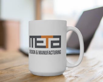 May include: White ceramic mug with a large handle. The mug features the word "META" in a black, gray, and orange block letter design, with the words "DESIGN & MANUFACTURING" printed below in gray.
