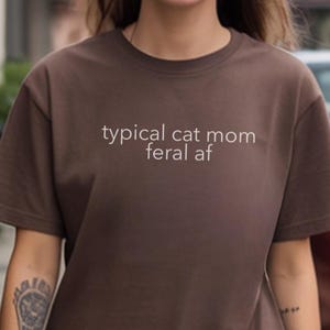Könnte beinhalten: Ein braunes T-Shirt mit dem weißen Text "typical cat mom feral af". Das Shirt hat einen Rundhalsausschnitt und kurze Ärmel. Das Bild ist im Freien aufgenommen.