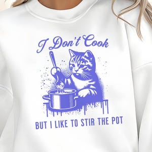 Puede incluir: Sudadera blanca con un gráfico azul de un gato removiendo una olla, con el texto "I Don't Cook BUT I LIKE TO STIR THE POT". La sudadera tiene cuello redondo y mangas largas. El gráfico está centrado en la parte delantera.