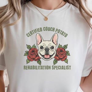 Könnte beinhalten: Ein weißes T-Shirt mit einer Grafik eines Französische Bulldogge-Gesichts, das zwischen zwei roten Rosen zentriert ist. Der Text "CERTIFIED COUCH POTATO" ist grün über dem Hund und "REHABILITATION SPECIALIST" ist grün unter dem Hund gedruckt.